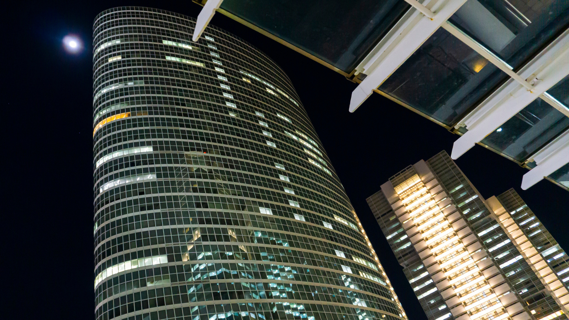 Inquadratura dal basso di un moderno grattacielo vetrato illuminato di notte a Tokyo sotto la luna, a simboleggiare la solidità della branch establishment e l'apertura di sedi aziendali.