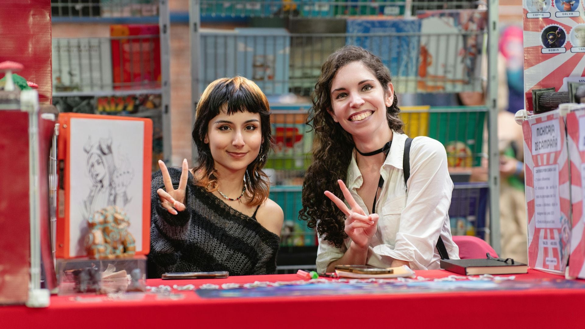 Due giovani ragazze sorridenti fanno il segno della pace dietro un banchetto di illustrazioni e merchandise durante un evento dedicato a manga e anime a Tokyo, rappresentando l'anima social e local della community.