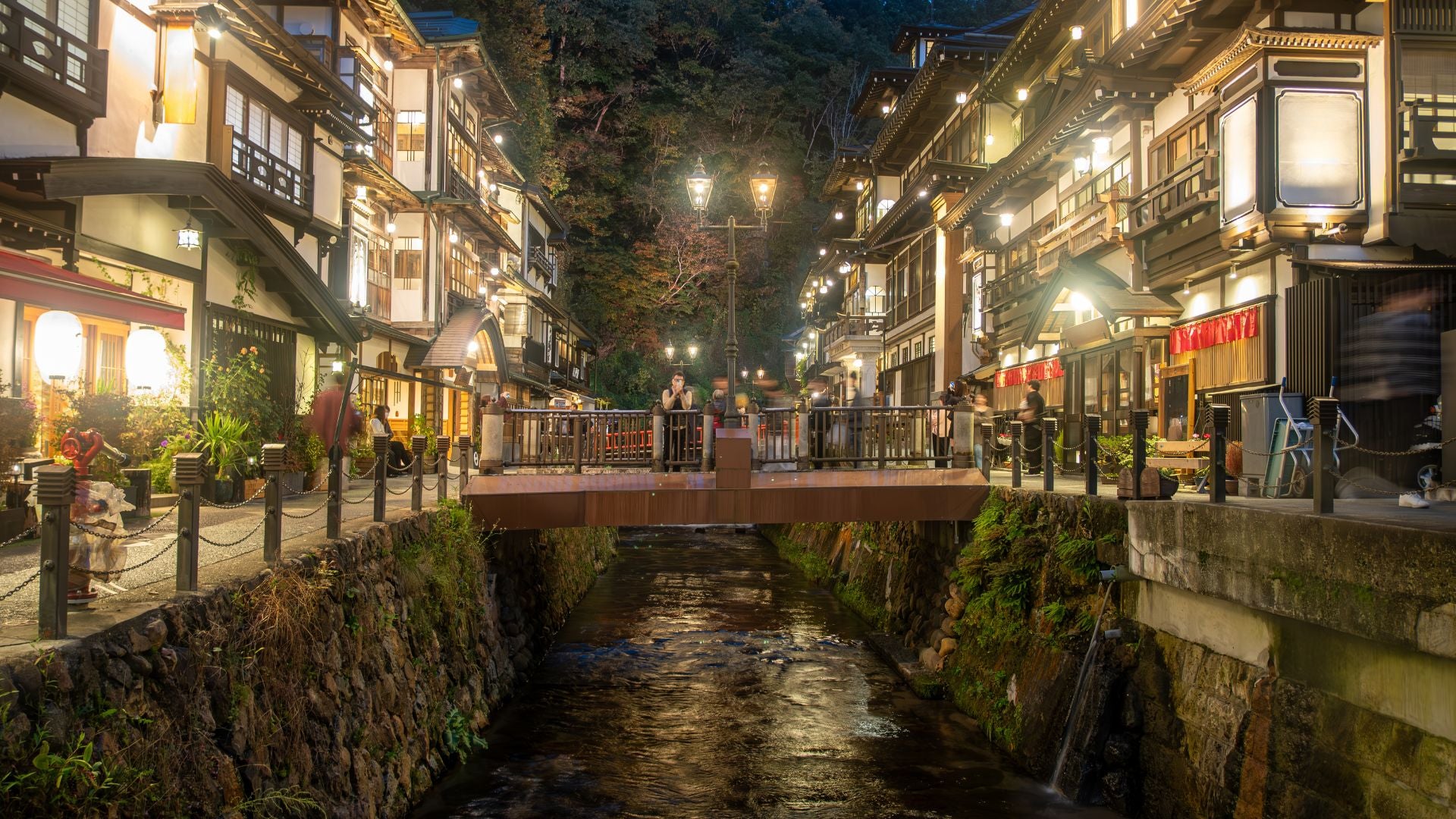 Suggestiva veduta notturna di Ginzan Onsen illuminata dalle lanterne calde che si riflettono nel torrente, evocando l'atmosfera magica e senza tempo del Giappone rurale.